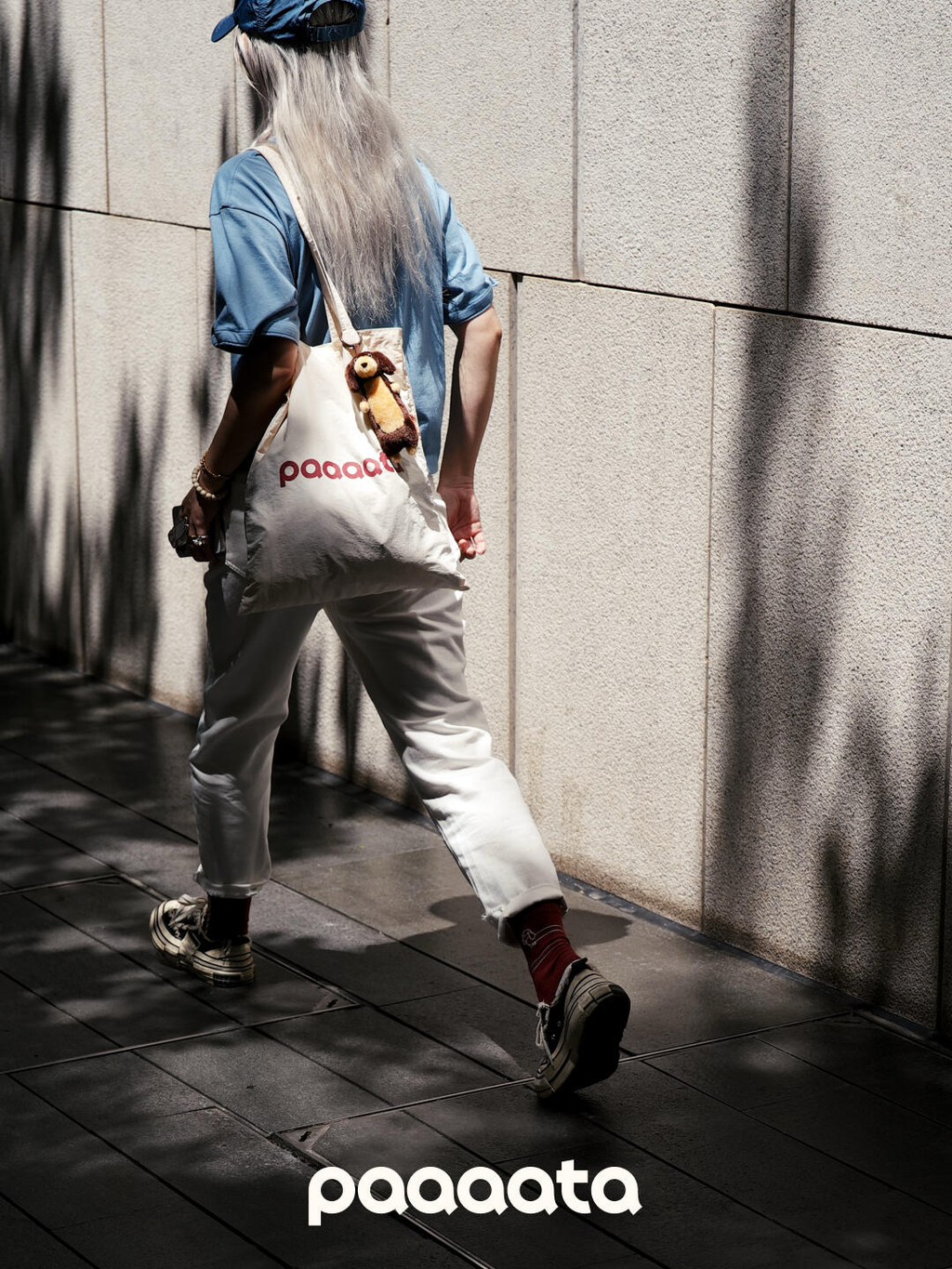 Person with a long white beard wearing a blue shirt and white pants, holding a white bag with 'paaaata' branding, against a textured wall.